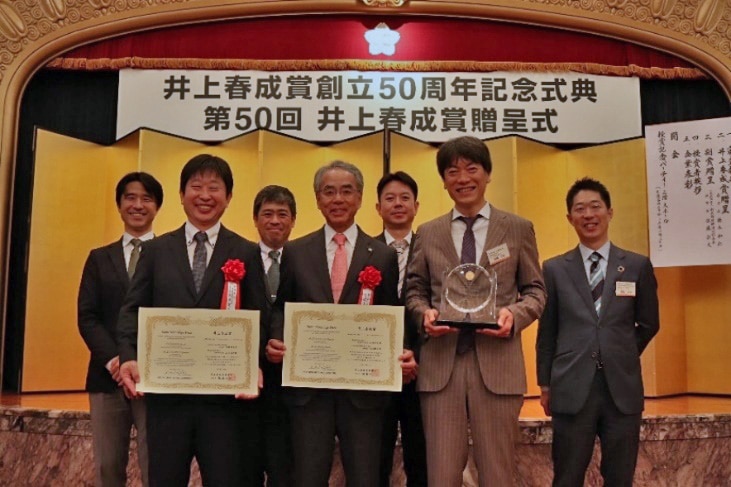 賞状を持つ当社代表取締役 山本靖則（前列中央）と 九州大学 生体防御医学研究所 馬場健史教授（前列左）