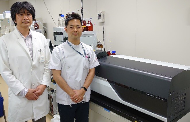 加藤隆弘准教授（左）、九州大学病院検査部の瀬戸山大樹助教 および研究で使用する高速液体クロマトグラフ質量分析計