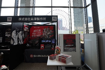 島津製作所ブース。モンスターの技術や番組本番までの裏話を記載したタブロイド紙や、ステッカーをプレゼントした